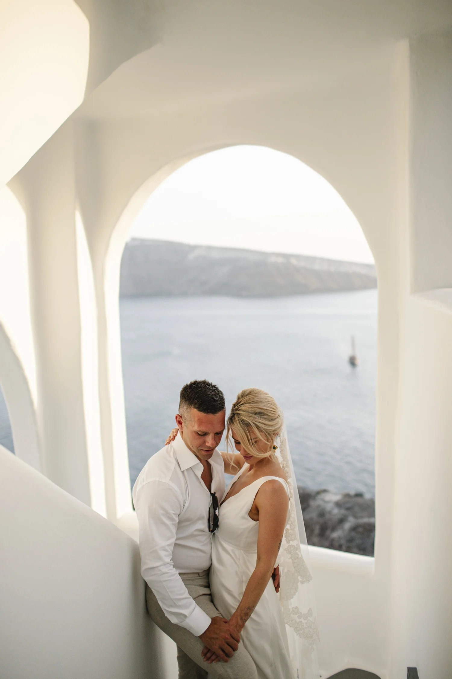 A couple dressed in wedding attire standing close together on a white structure with a scenic ocean view in the background. The man is wearing a white shirt and light gray pants, and the woman is in a white wedding dress with lace details. They are holding hands and looking down, with their foreheads touching, during a romantic moment.