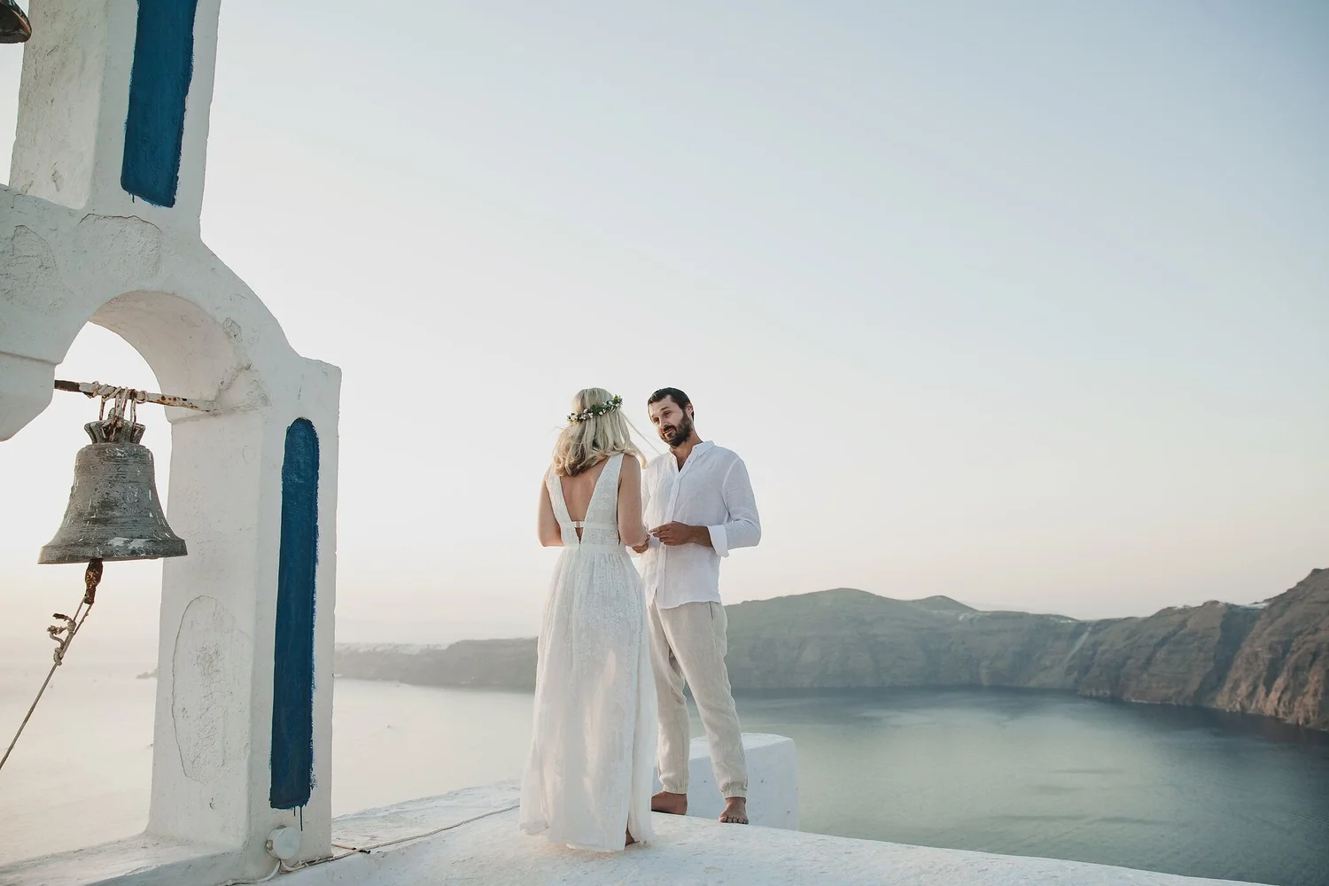 A couple dressed in white standing together near a white bell tower with a mountain and water background at sunset.