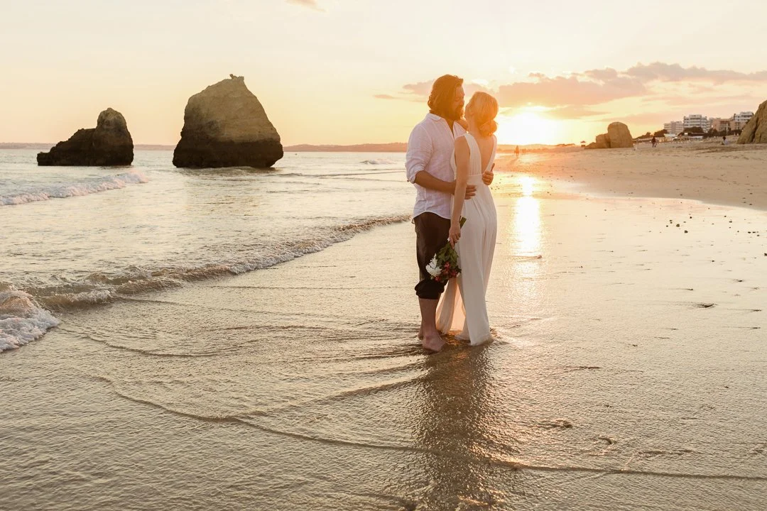 A couple standing arm in arm in shallow water on a beach at sunset, with rocks and buildings in the background.