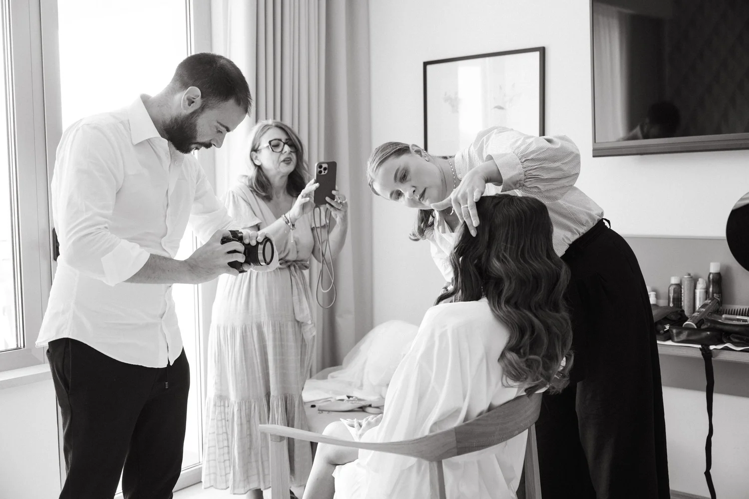 A woman hairstylist styling a woman's hair in a salon, while a man is taking a photo with a camera and a woman in the background is holding a phone.