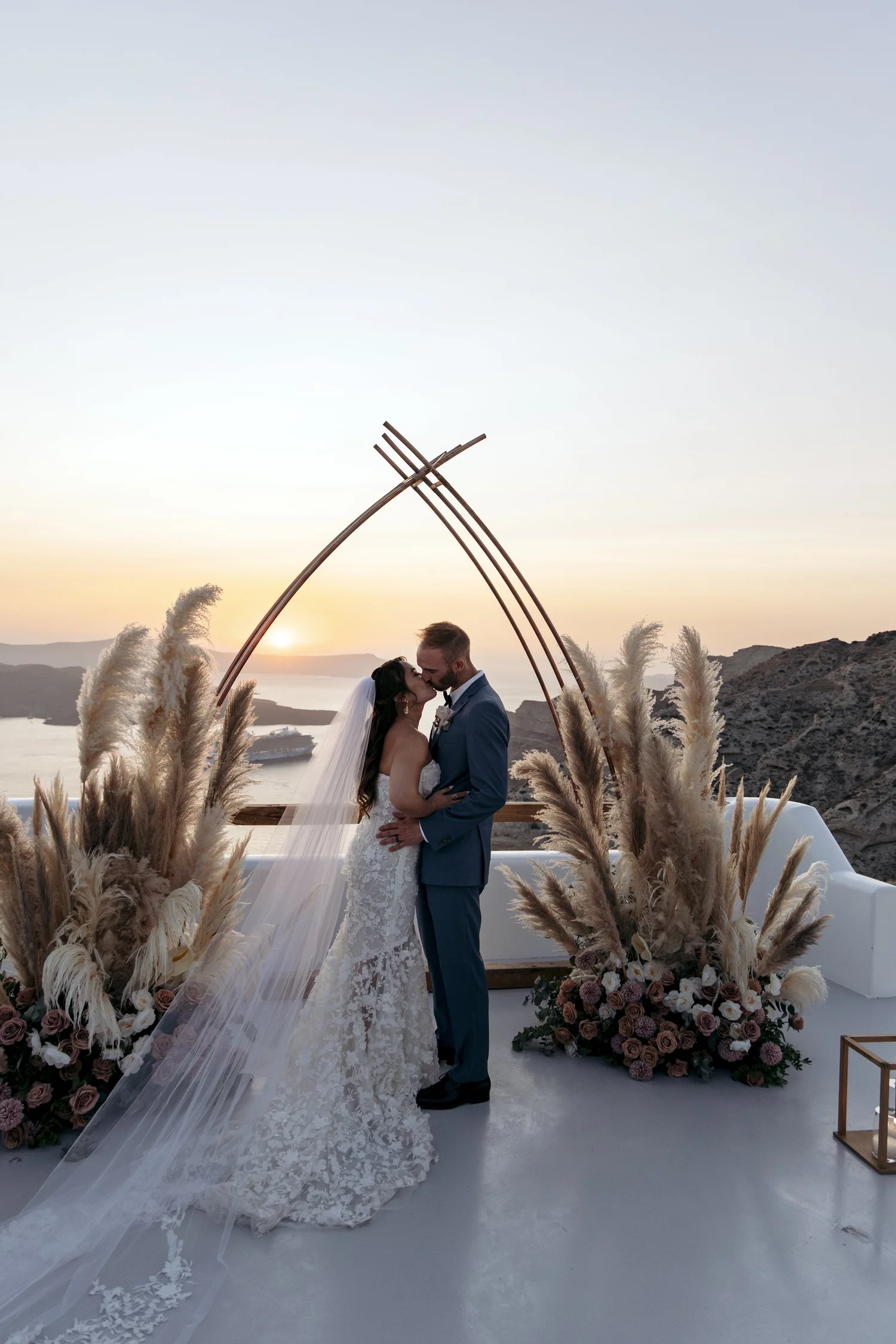 A bride and groom sharing a kiss during their wedding ceremony at sunset, with floral arrangements, pampas grass, and a scenic landscape in the background.