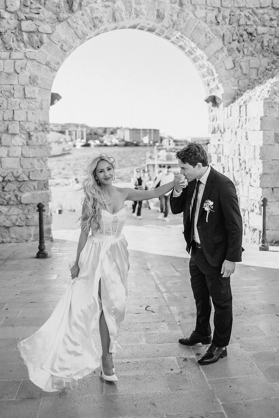 A bride and groom are posing beneath a stone arch, with the bride touching the groom's hand to her lips. The bride is wearing a white wedding dress and high heels, while the groom is in a dark suit with a boutonnière. The background shows a waterfron