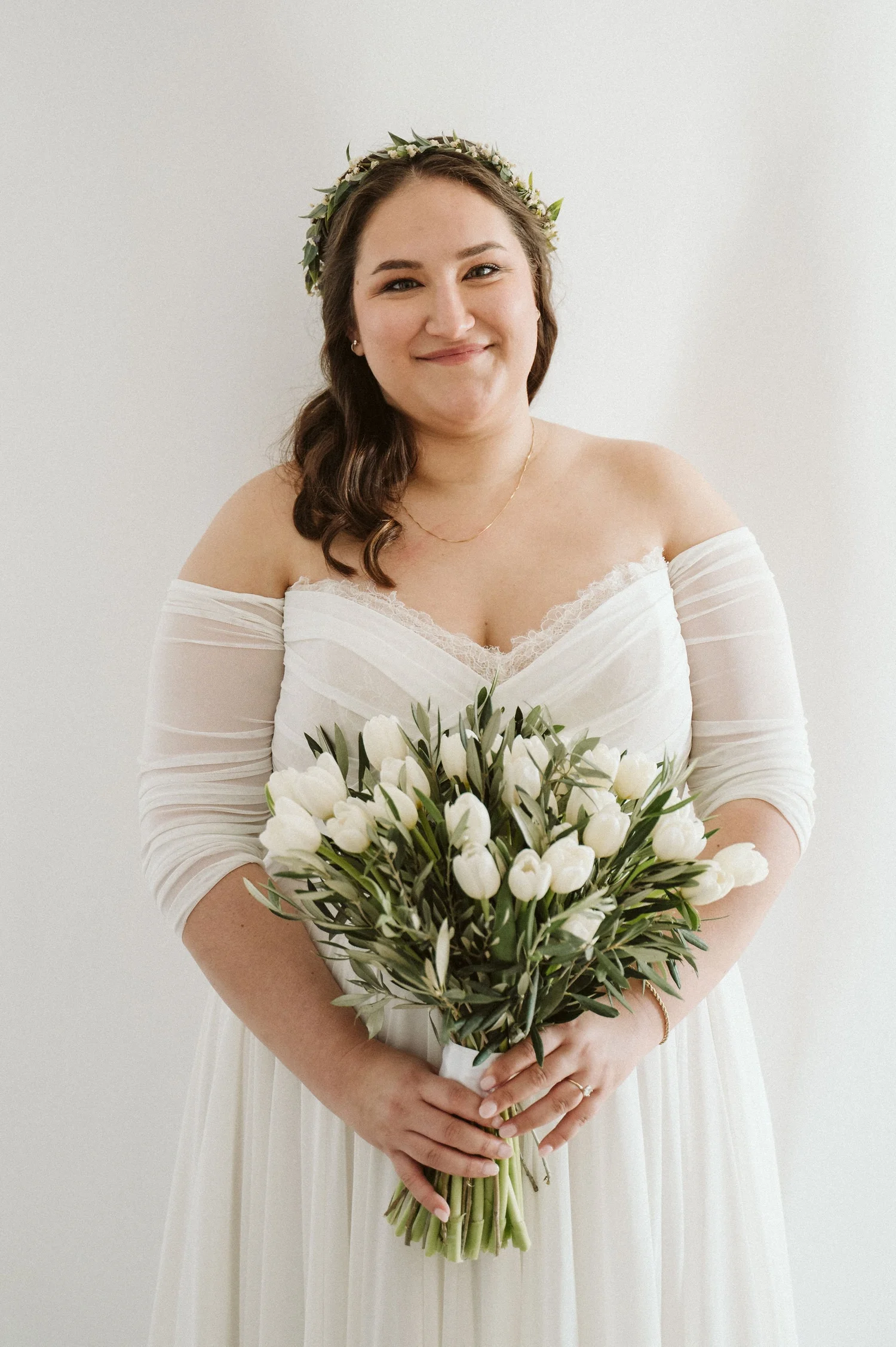A smiling woman dressed in a white off-the-shoulder wedding gown, wearing a floral crown, holding a bouquet of white tulips with green leaves, in front of a plain white background.