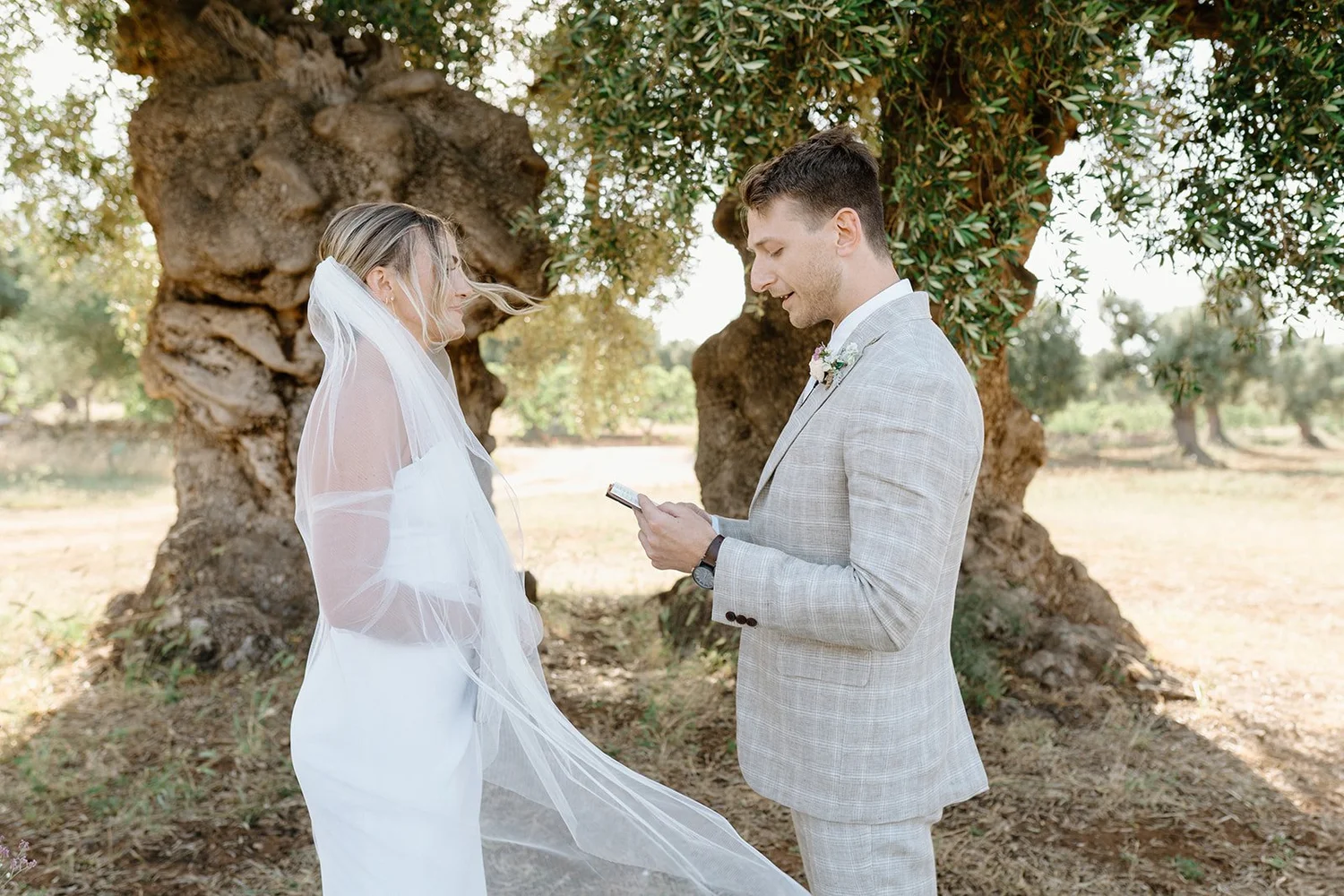 A couple stands outdoors in front of a large tree during their wedding ceremony. The bride wears a white dress and veil, and the groom wears a light gray checked suit with a white shirt. The groom is reading vows from his phone, and they are smiling