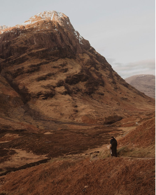 A person standing on grassy terrain in a valley surrounded by mountains, with a snow-capped peak in the distance.