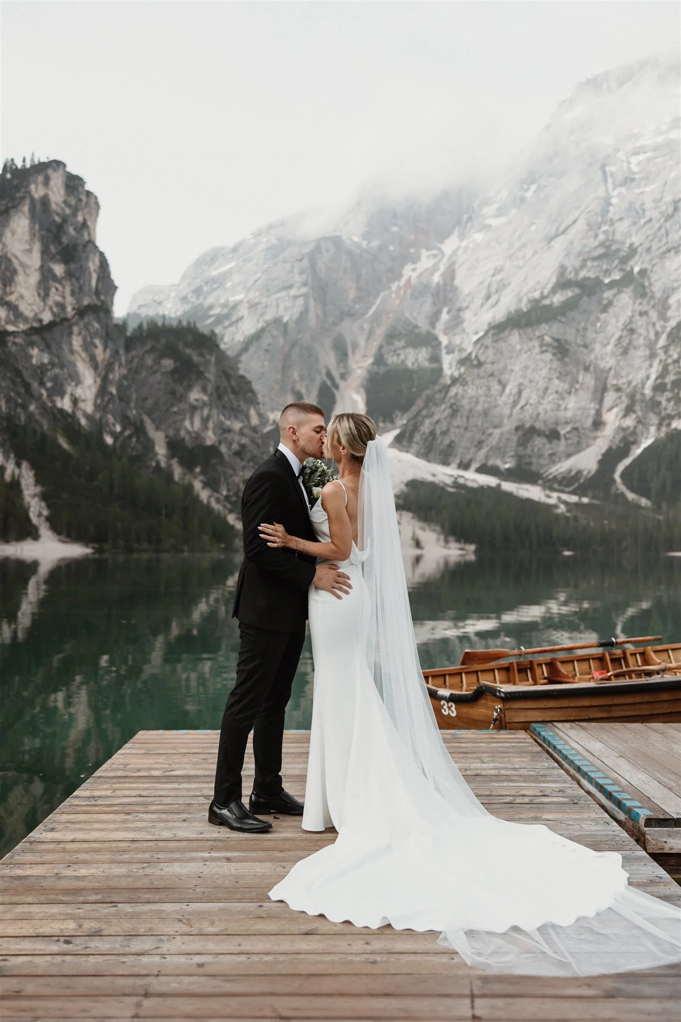 A bride and groom embrace and kiss on a wooden dock beside a mountain lake, with snow-capped mountains and misty skies in the background. The bride wears a white dress with a long train and veil, while the groom wears a black tuxedo. A small wooden boat is moored to the dock.