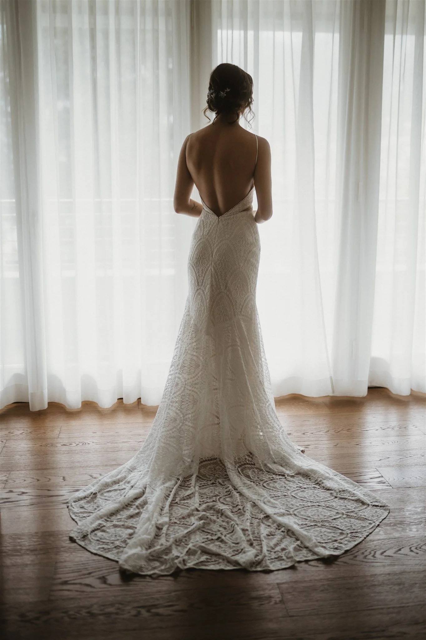 A bride standing indoors in front of sheer curtains, wearing a backless, lace wedding gown with a long train, holding her arms behind her back.