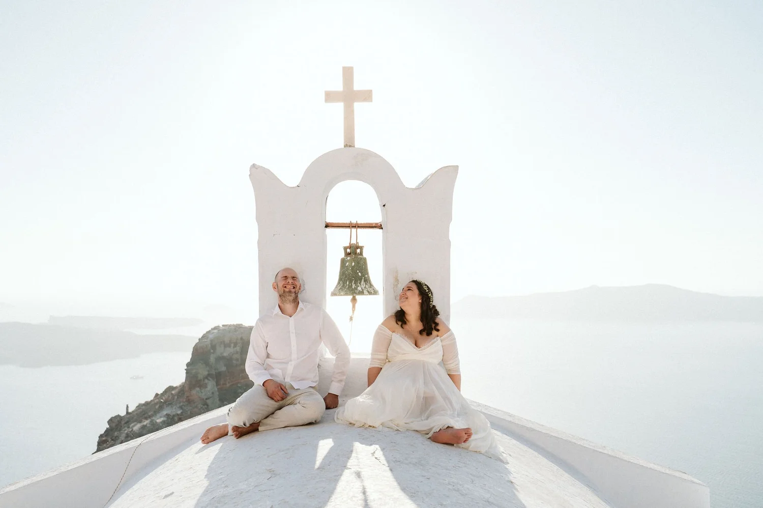 A couple dressed in white sitting on the roof of a white church with a bell and cross in a snowy landscape.