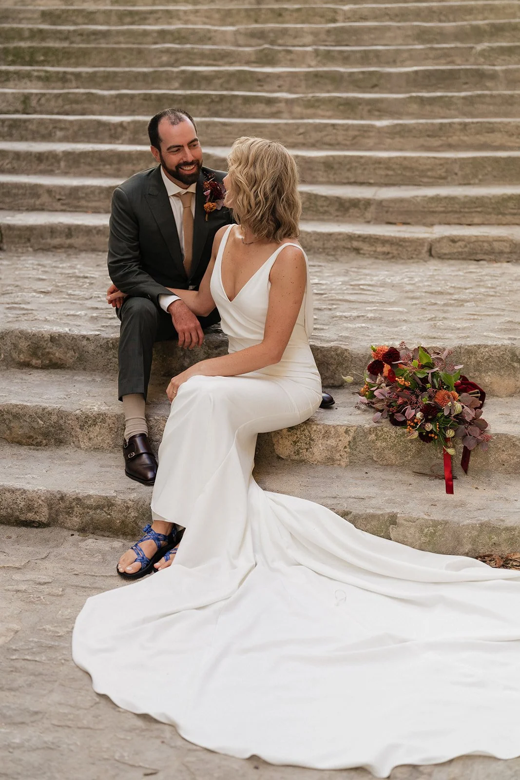 A bride and groom sitting on stone steps, gazing at each other, with a bouquet of flowers on the ground beside them.
