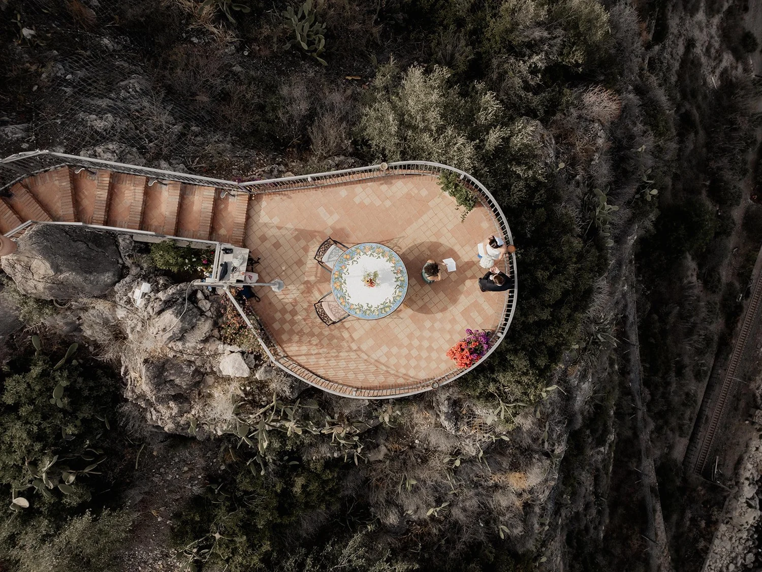An aerial view of a small outdoor terrace with a round table and four chairs, situated on a rocky hillside with surrounding desert vegetation. Three people are standing near the table, engaged in conversation. The terrace has a tiled floor and a curv