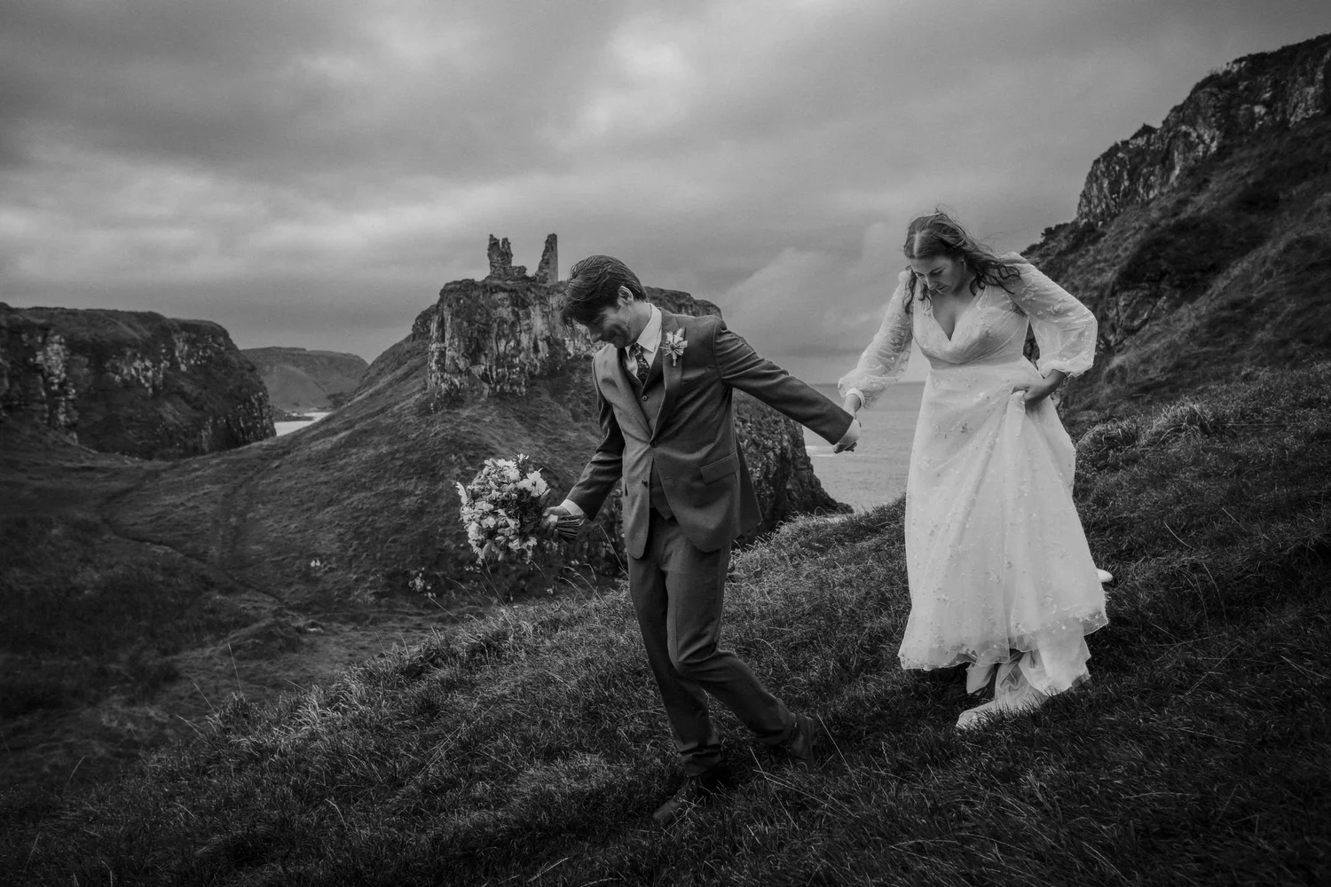 A black-and-white photo of a bride and groom holding hands and walking on a grassy hillside, with rocky cliffs and a cloudy sky in the background.