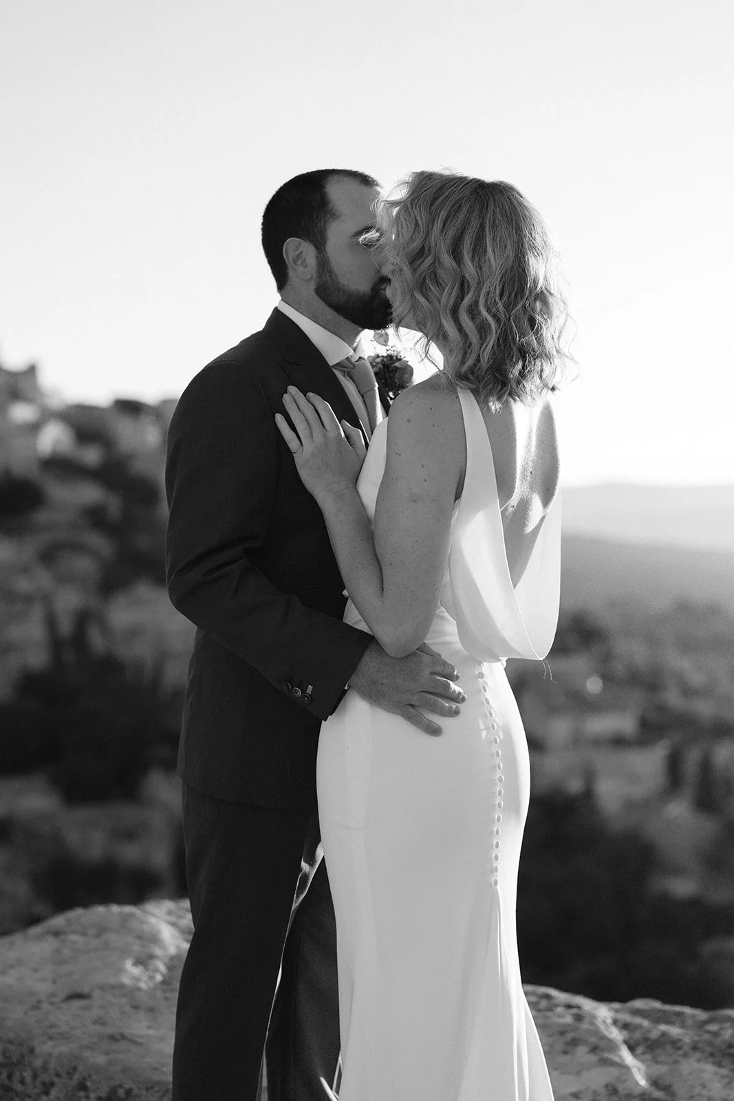 A black and white photo of a couple sharing a kiss outdoors, the man in a suit and the woman in a sleeveless wedding dress with buttons down the back.