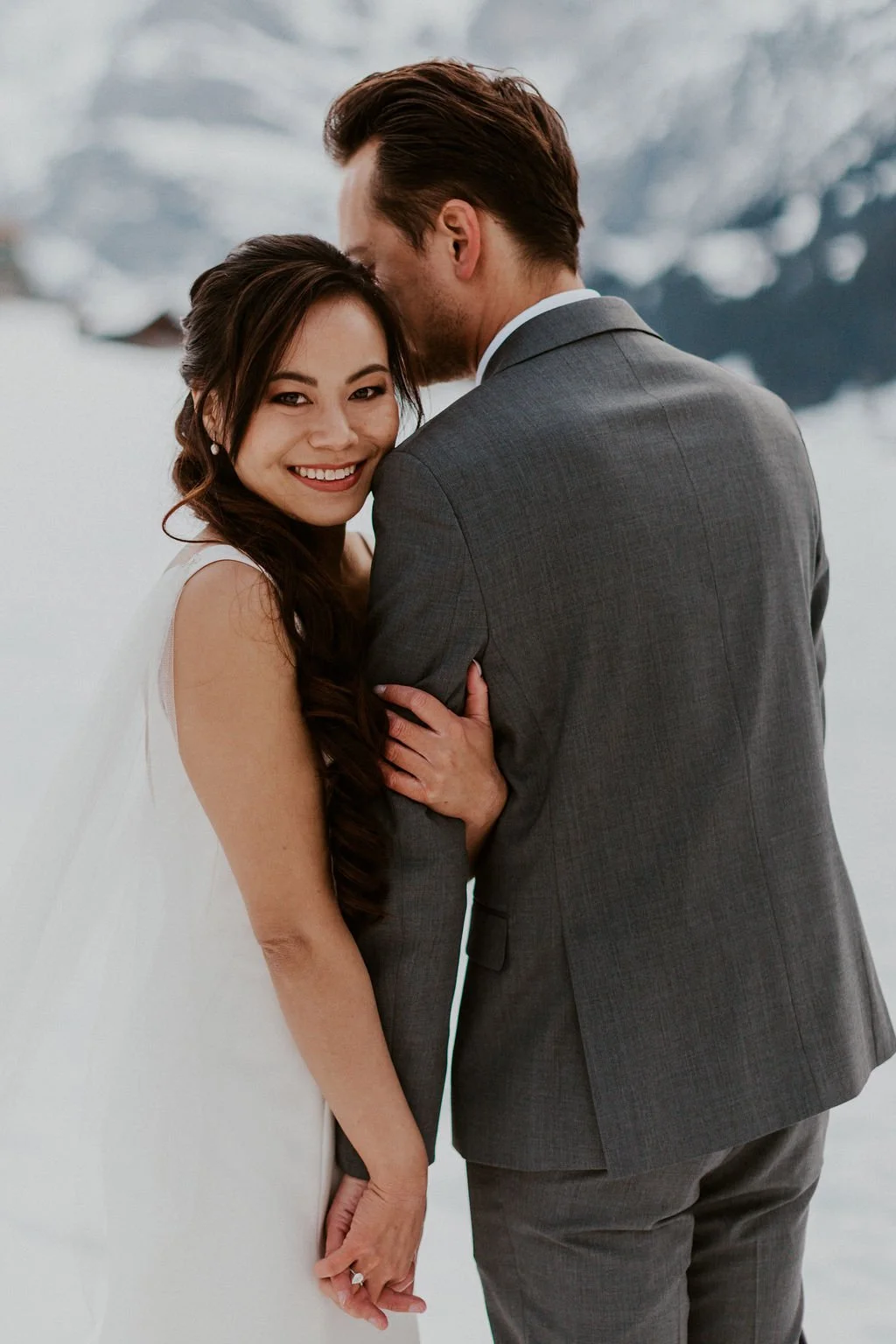 A bride and groom embrace outdoors in a snowy landscape, with the bride smiling at the camera and the groom with his face close to her forehead.