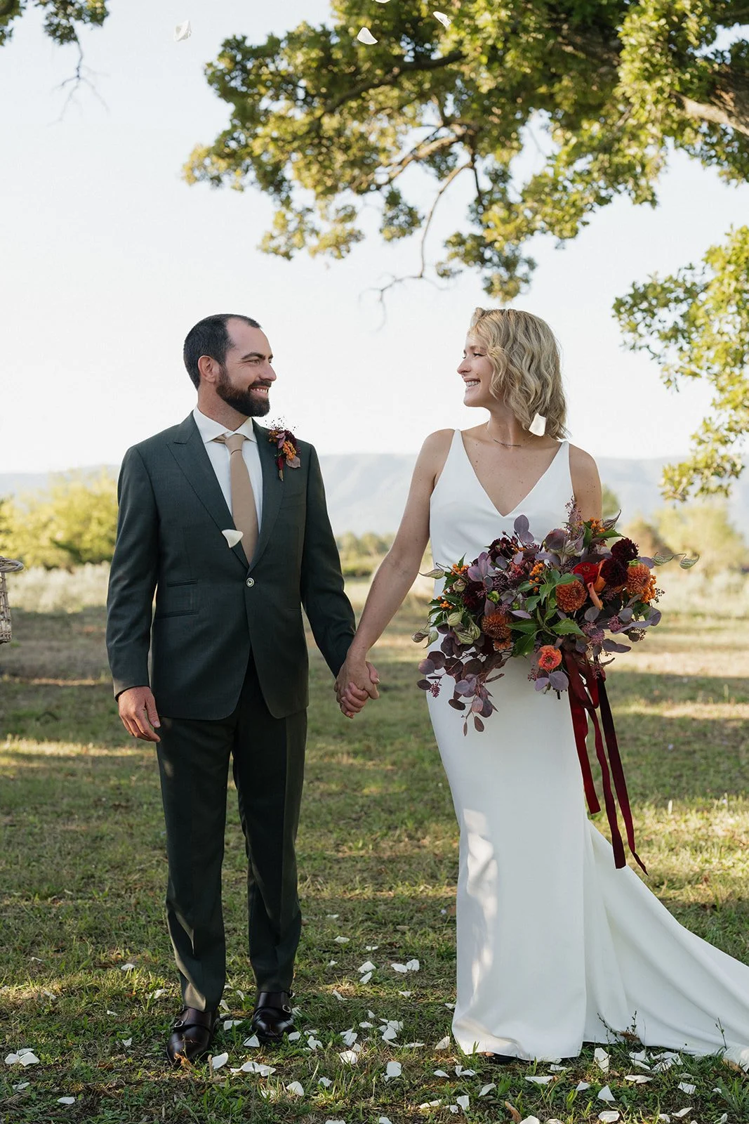 A bride and groom holding hands outdoors, smiling at each other, with trees and distant mountains in the background. The bride wears a white gown and holds a large bouquet, and the groom wears a dark suit with a tie.
