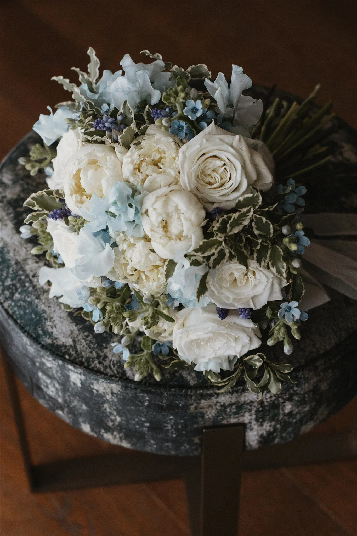A bouquet of white roses, light blue and purple flowers with variegated leaves, placed on a dark granite table.