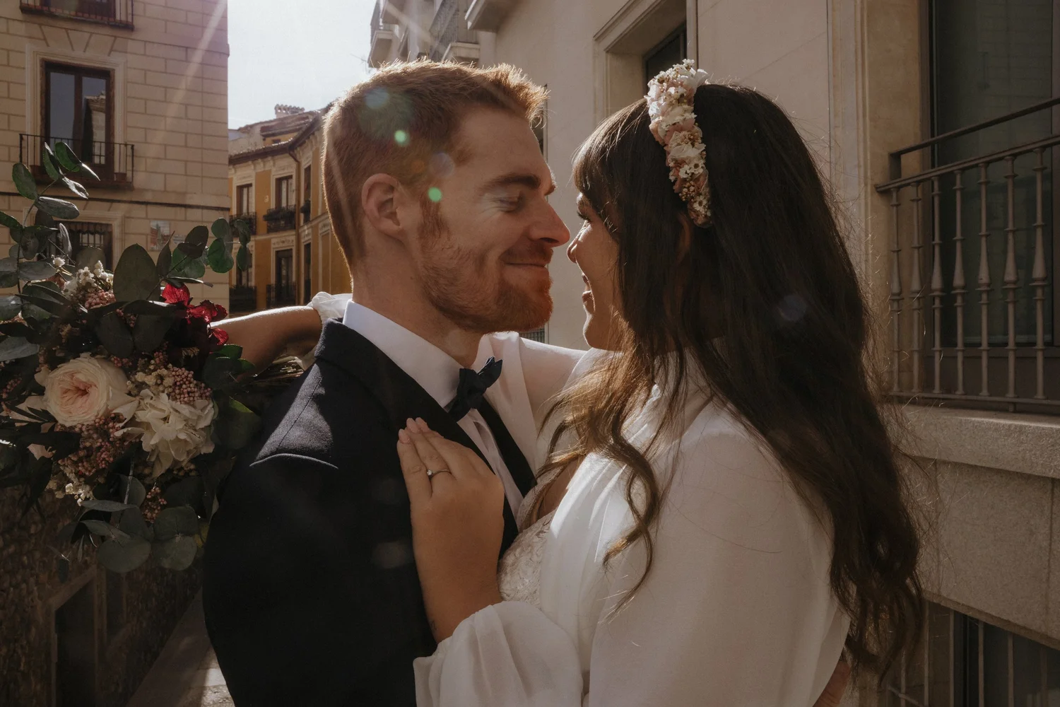 A couple dressed in wedding attire, embracing and smiling on a city street during daytime.