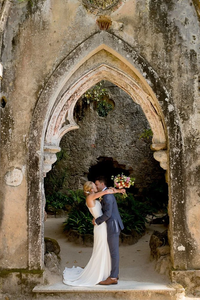 A bride and groom kissing under an arched stone doorway with plants and rocks in the background.