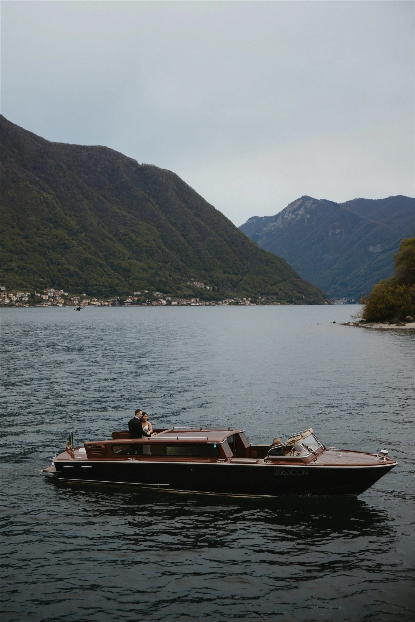 A couple on a yacht with two people sitting on the deck, sailing on a body of water with mountain scenery in the background.
