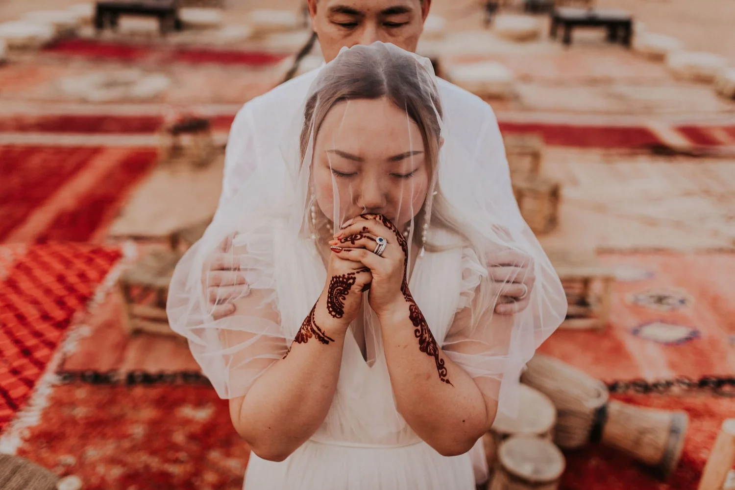 A bride praying in a wedding ceremony with henna designs on her hands, wearing a white dress and veil, standing in front of a person, in a room with red patterned carpets.