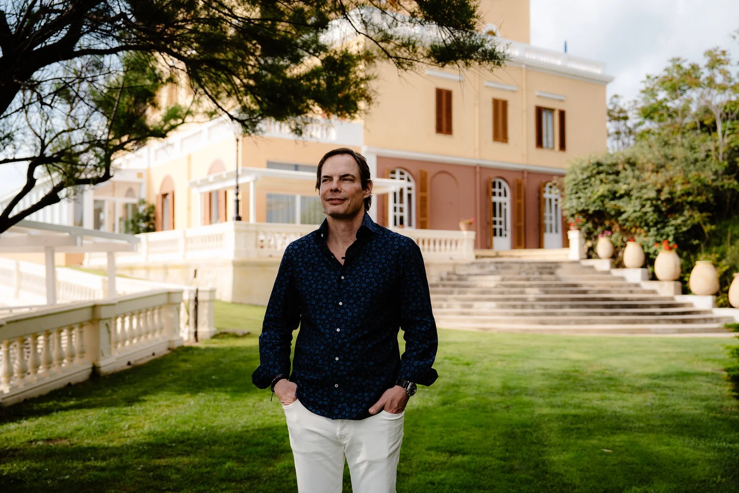A man with dark hair wearing a dark patterned shirt and white pants standing outside near a yellow building with brown shutters and stairs, with trees and greenery around.