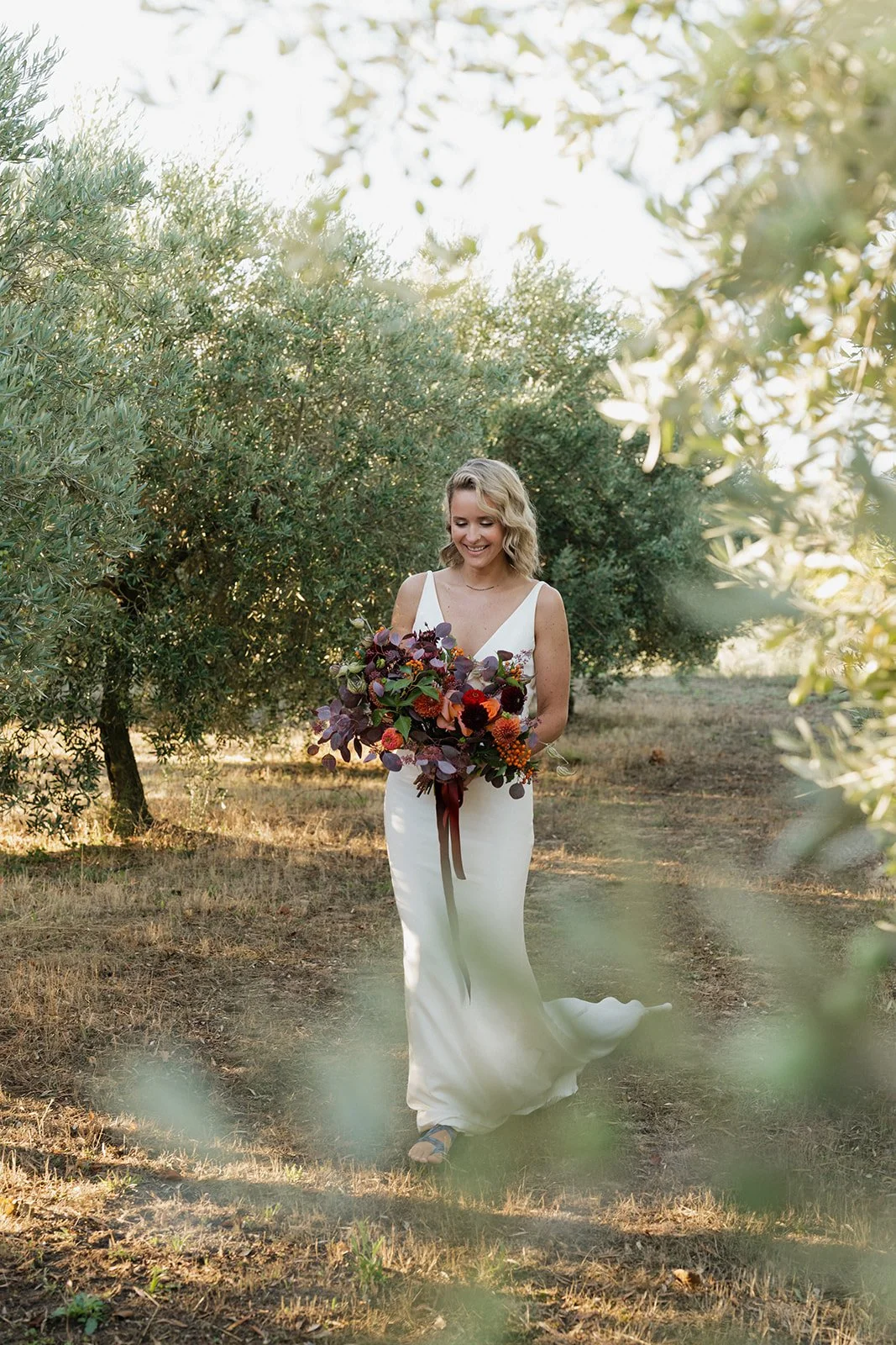 A smiling woman in a white dress walking outdoors through an olive grove, holding a large bouquet of dark and reddish flowers with greenery.