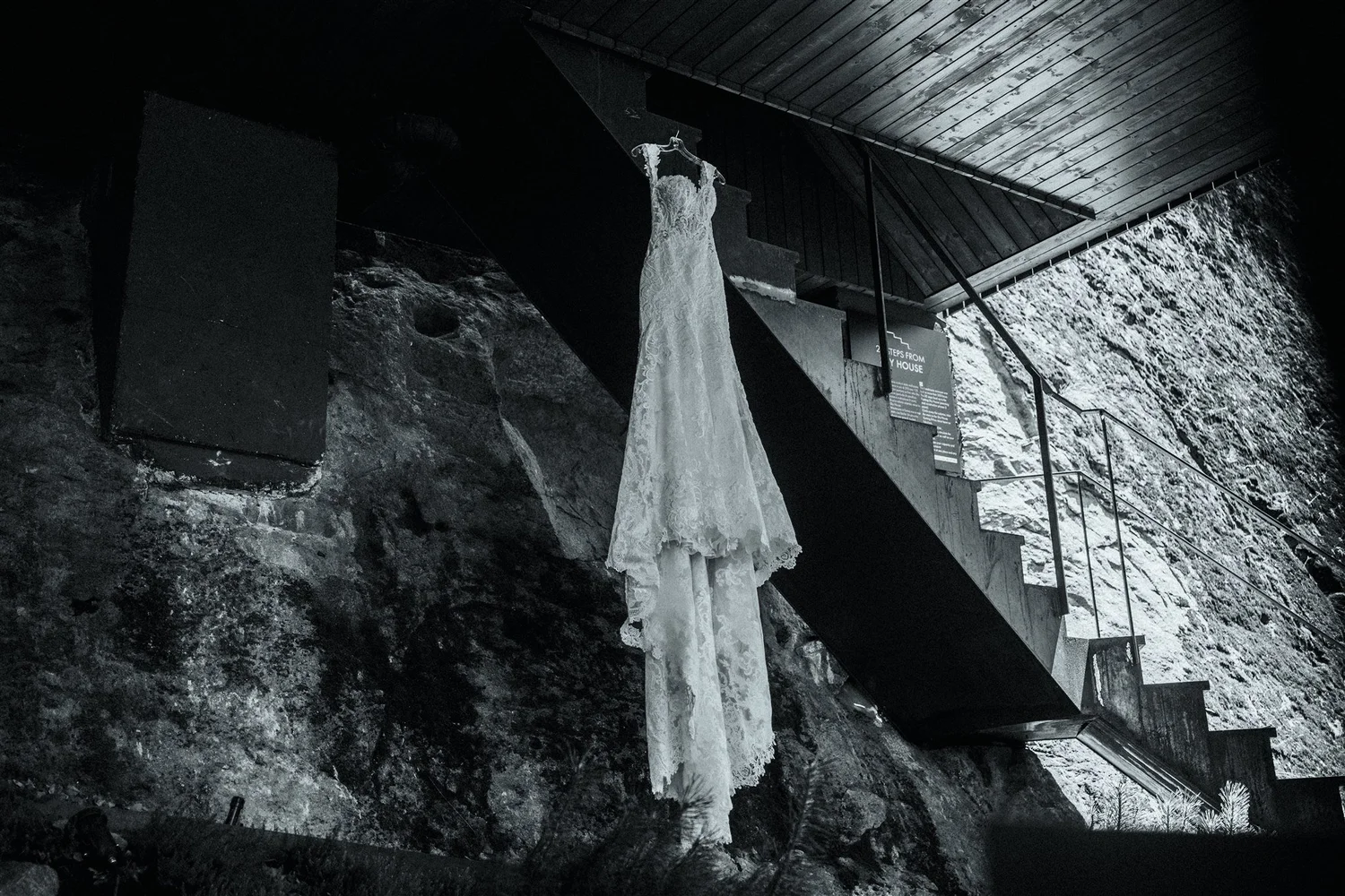 A white wedding dress hanging on a hanger from a railing in a dark, rocky setting, with a staircase and informational signs nearby.
