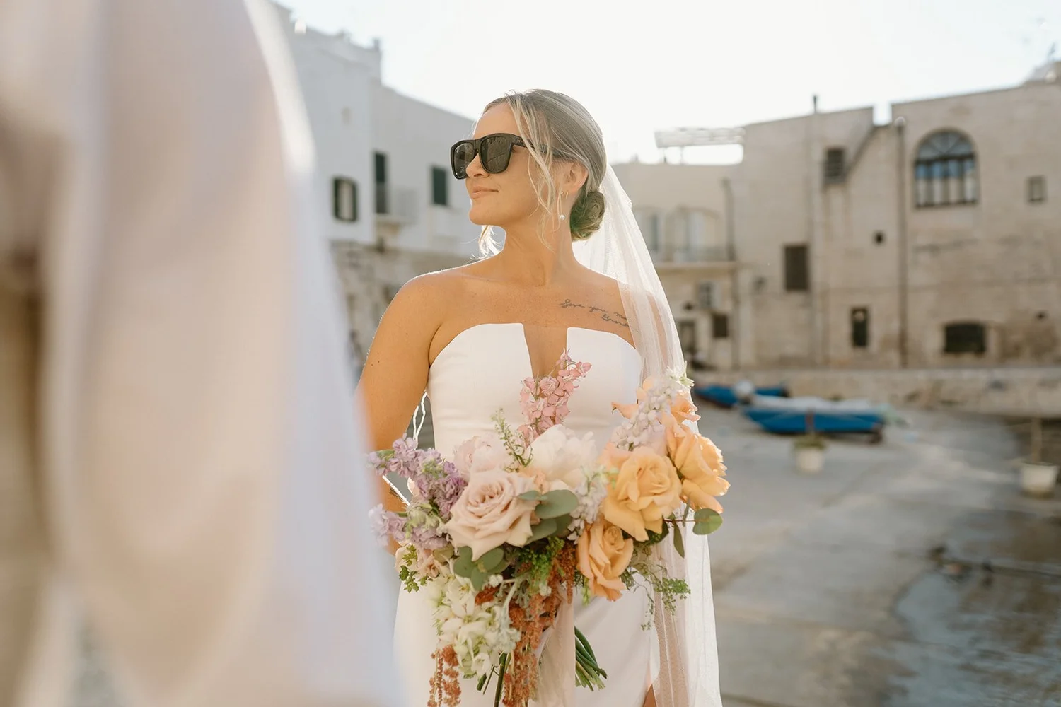 A bride holding a bouquet of pink, beige, and white flowers, wearing a strapless white wedding dress and sunglasses, standing outdoors near a seaside with old buildings and boats in the background.