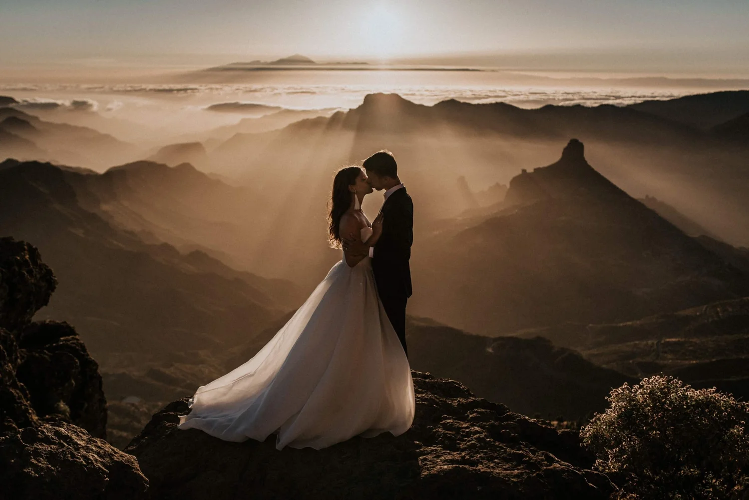 A bride and groom standing close together on rocky terrain, sharing a kiss with a mountainous landscape and clouds in the background during sunset or sunrise.