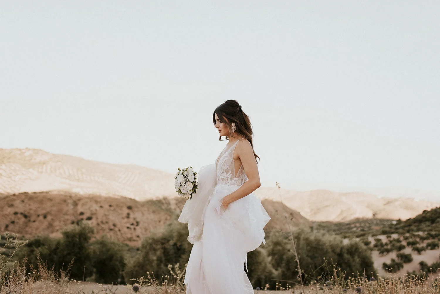 A bride in a white wedding dress holding a bouquet, standing outdoors in a desert landscape with mountains in the background.