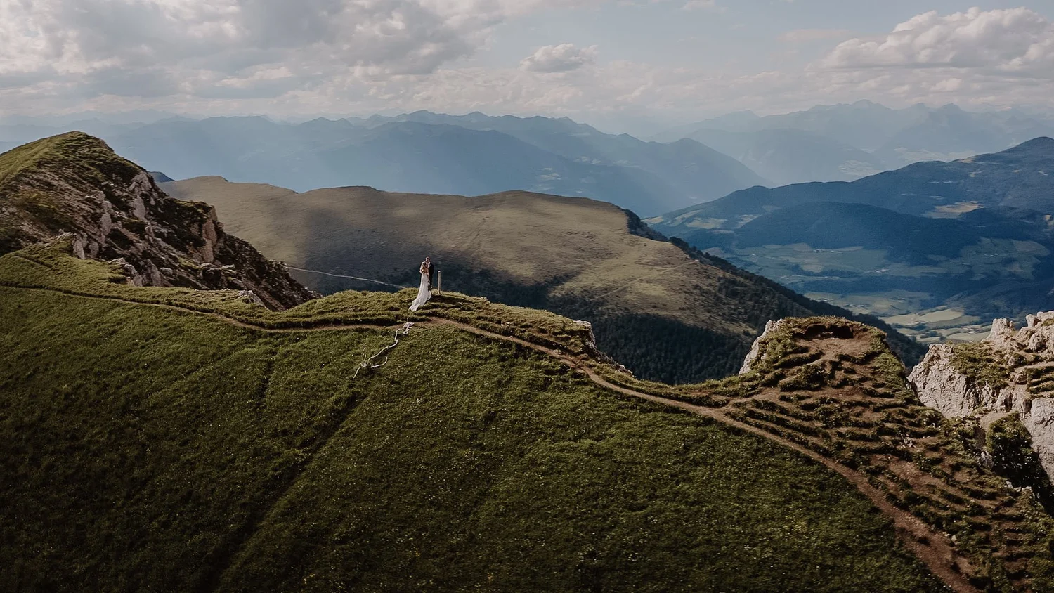 A couple in wedding attire standing on a grassy mountain ridge with a scenic view of rolling hills and distant mountains under a partly cloudy sky.