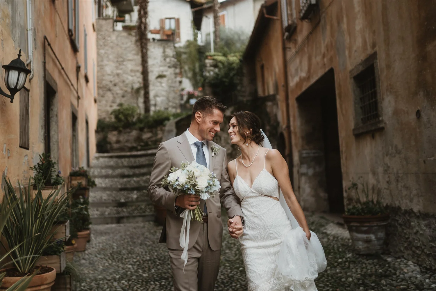 A bride and groom walking hand in hand on a cobblestone street in a rustic European village, smiling and holding hands. The groom is in a beige suit with a blue tie and boutonniere, holding a bouquet of white flowers. The bride is in a white lace wed
