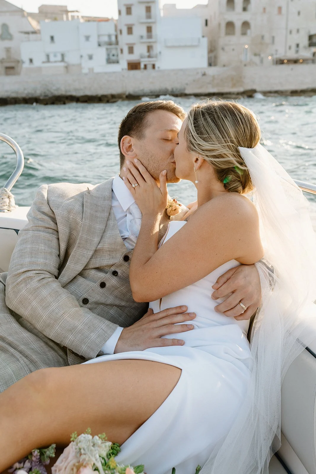 A newlywed couple sharing a kiss on a boat with water and buildings in the background.