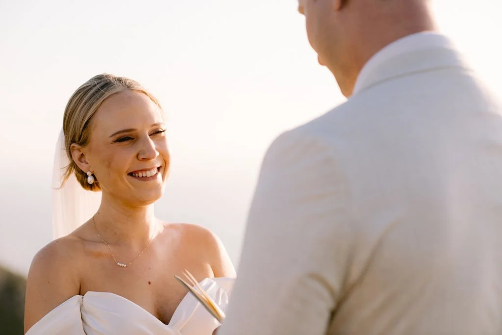 A bride smiling during her wedding ceremony outdoors, wearing a white wedding dress and jewelry, with a groom in a white shirt facing her.