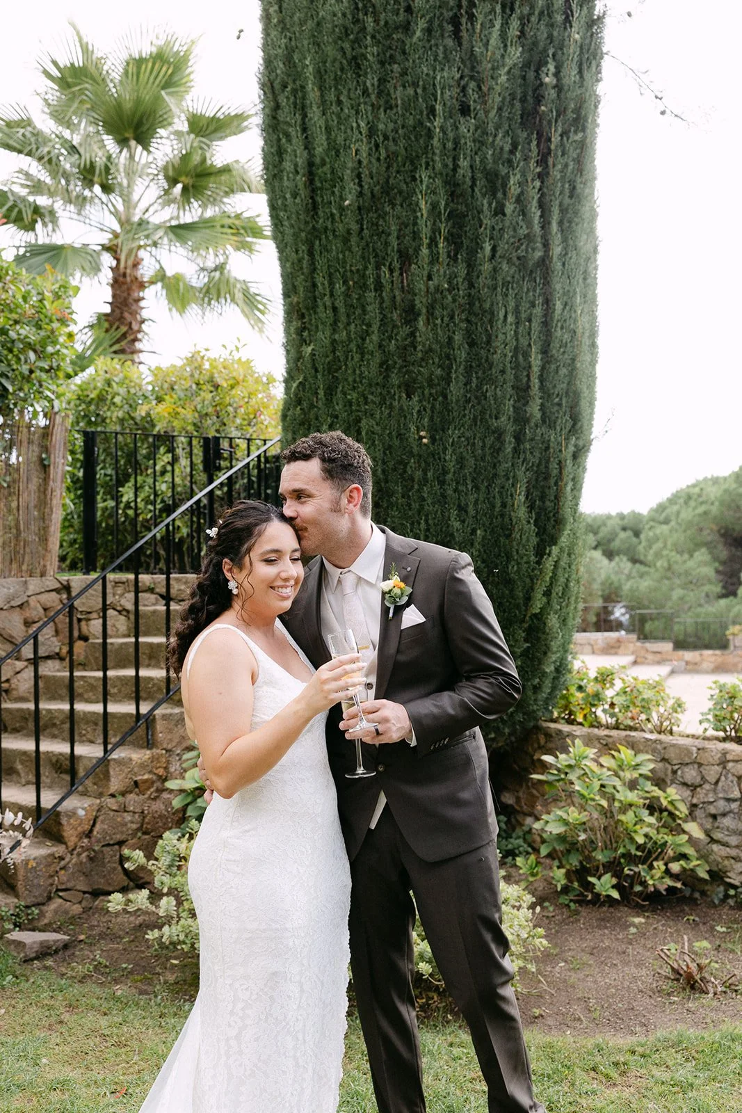 A newlywed couple celebrating outdoors, woman in a white lace wedding gown with dark curly hair, man in a dark suit with a boutonniere, holding a champagne flute, embracing near a stone staircase and lush greenery.