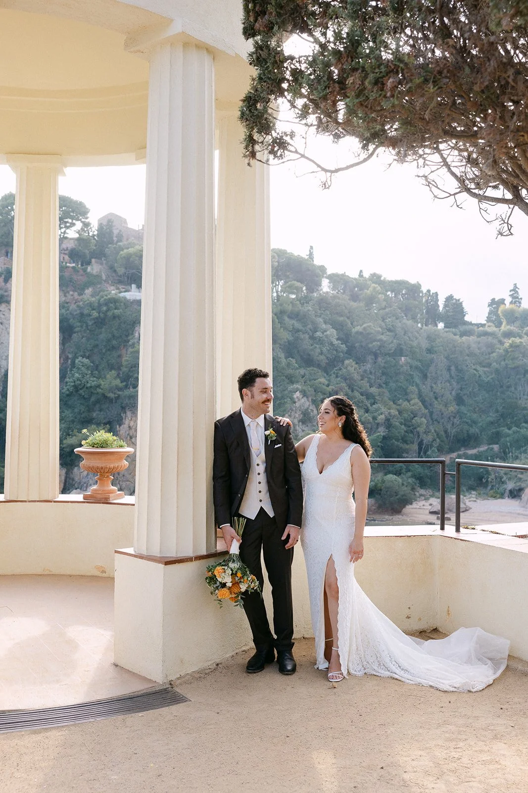 A bride and groom in wedding attire standing outdoors near large classical columns with a scenic hillside and trees in the background, smiling at each other.
