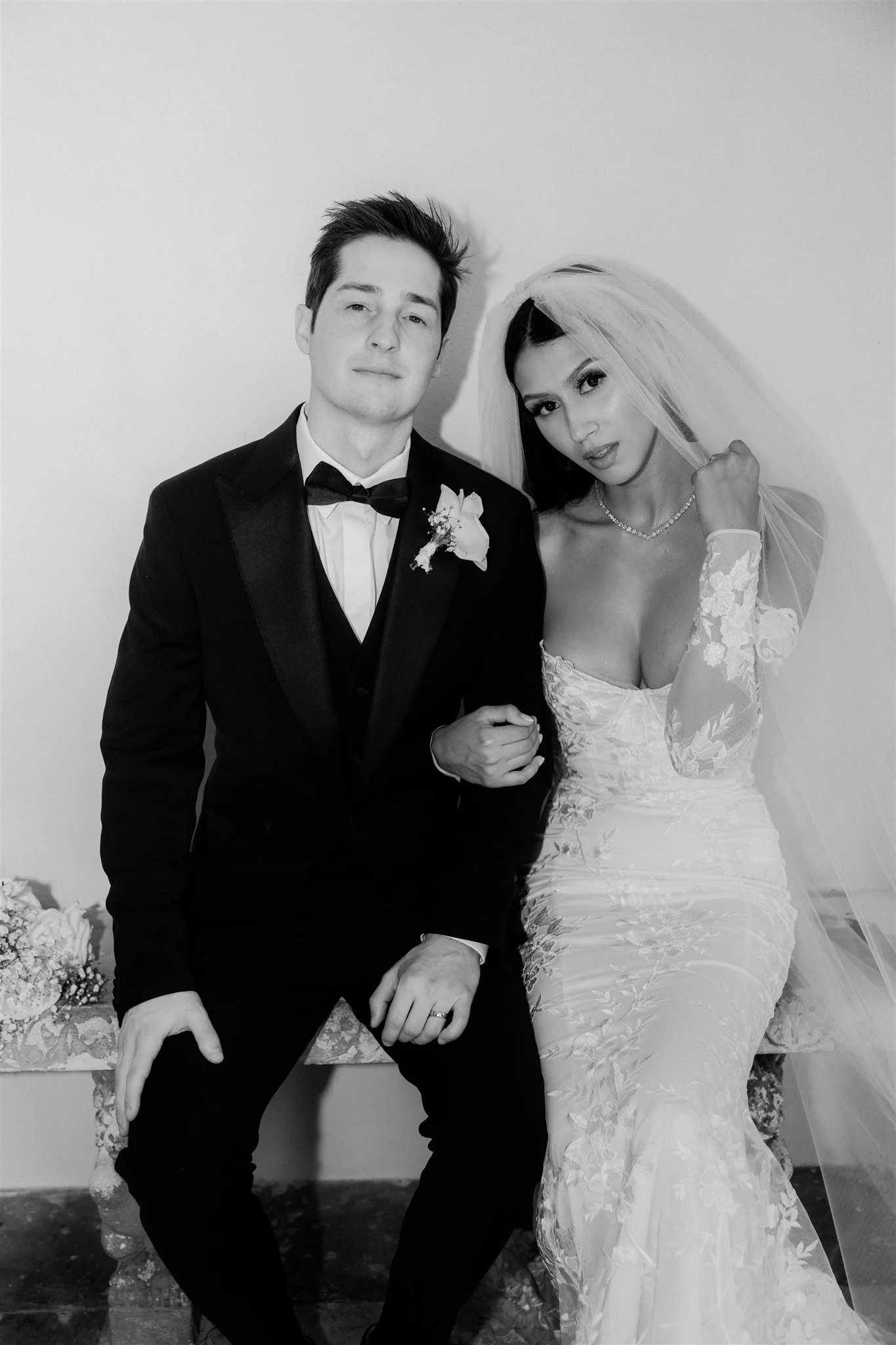 A black and white photo of a bride and groom. The groom is sitting, wearing a tuxedo with a bow tie and a boutonniere. The bride is standing beside him, wearing a lace wedding dress and a veil, holding his hand, and looking at the camera.