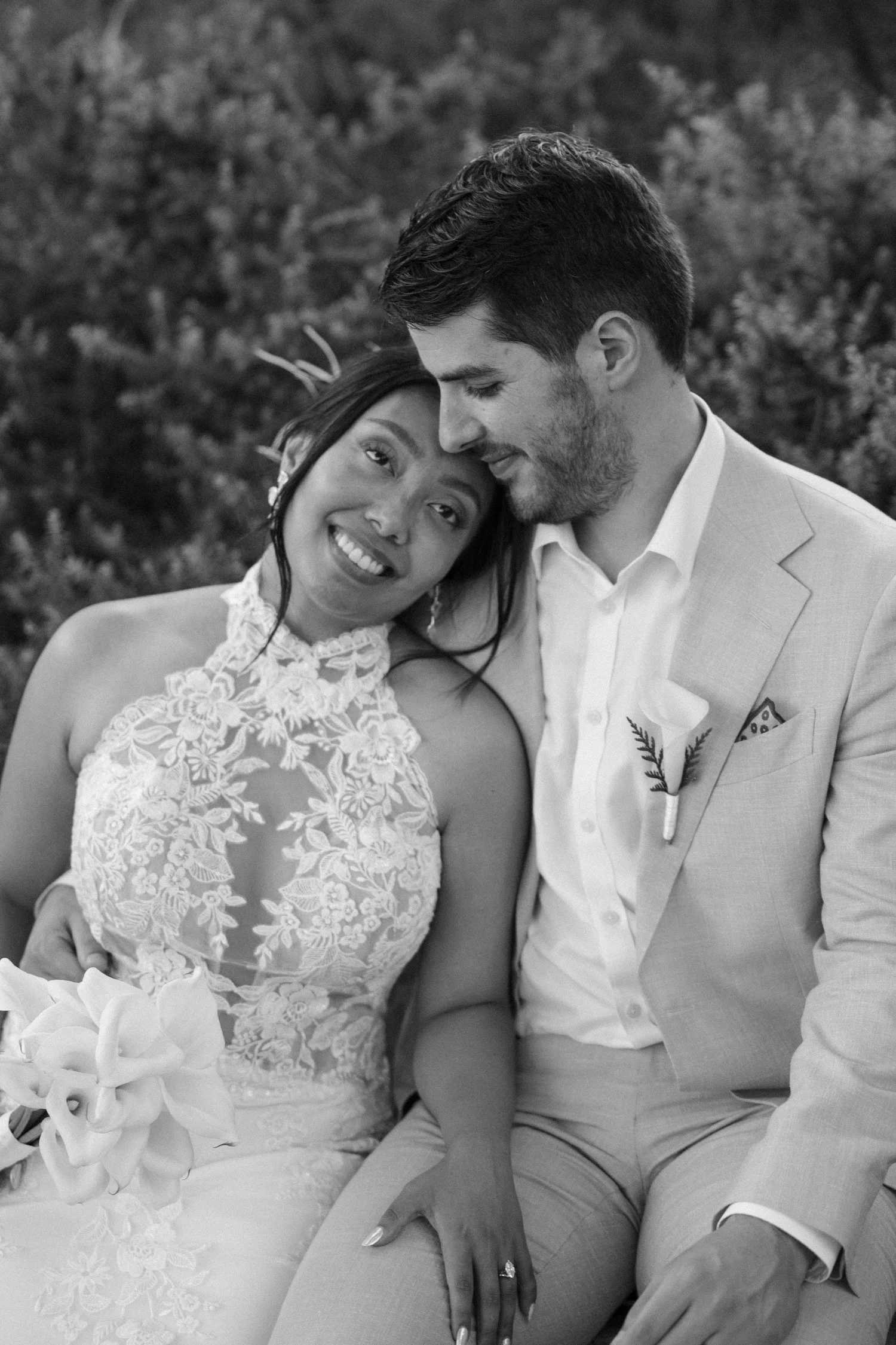 A black-and-white photo of a newlywed couple sitting closely together outdoors, with the woman wearing a lacy wedding dress and the man in a light-colored suit. They are smiling softly and leaning their foreheads together, with flowers in the foregro