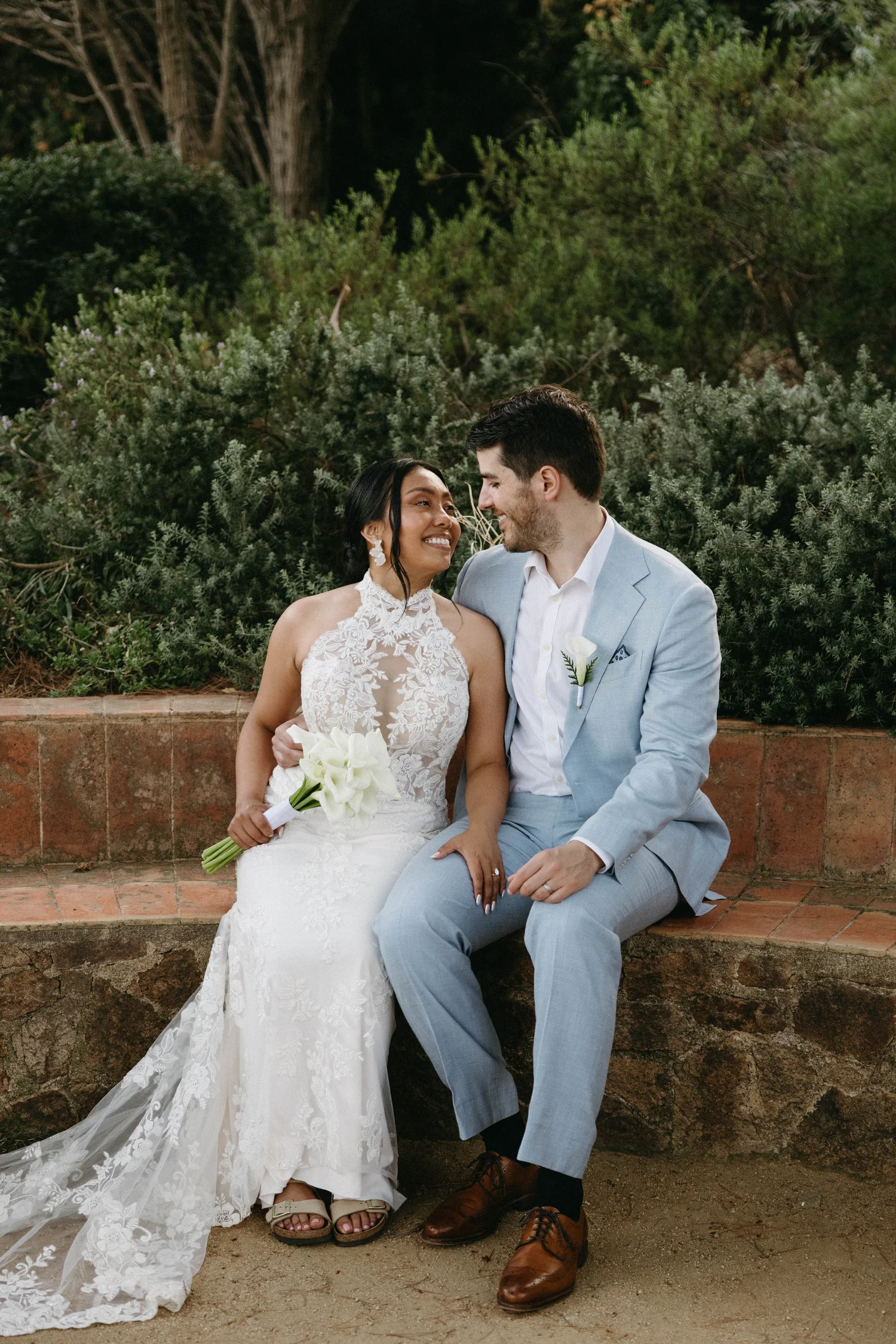 A bride and groom sitting on a brick ledge, smiling and looking at each other, outdoors surrounded by green bushes and trees, during their wedding celebration.