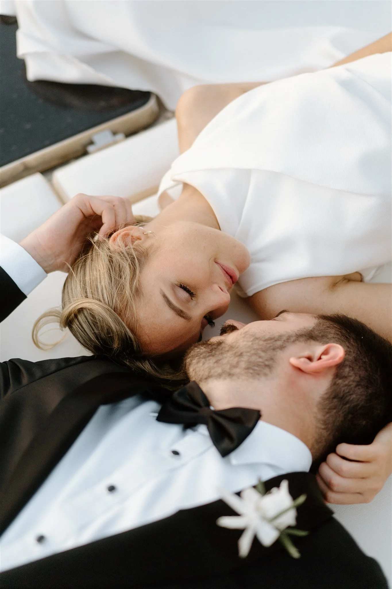 A couple lying on the floor, smiling and close to each other, dressed in formal wedding attire, with the woman wearing a white dress and the man in a tuxedo with a bow tie, celebrating their wedding day.