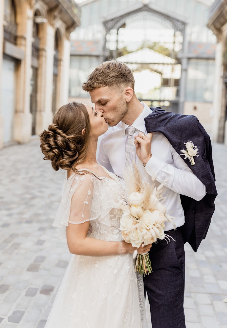 A newlywed couple sharing a kiss outdoors on a sunny day, with the bride holding a bouquet of white roses and the groom wearing a white shirt and dark suit jacket draped over his shoulder.