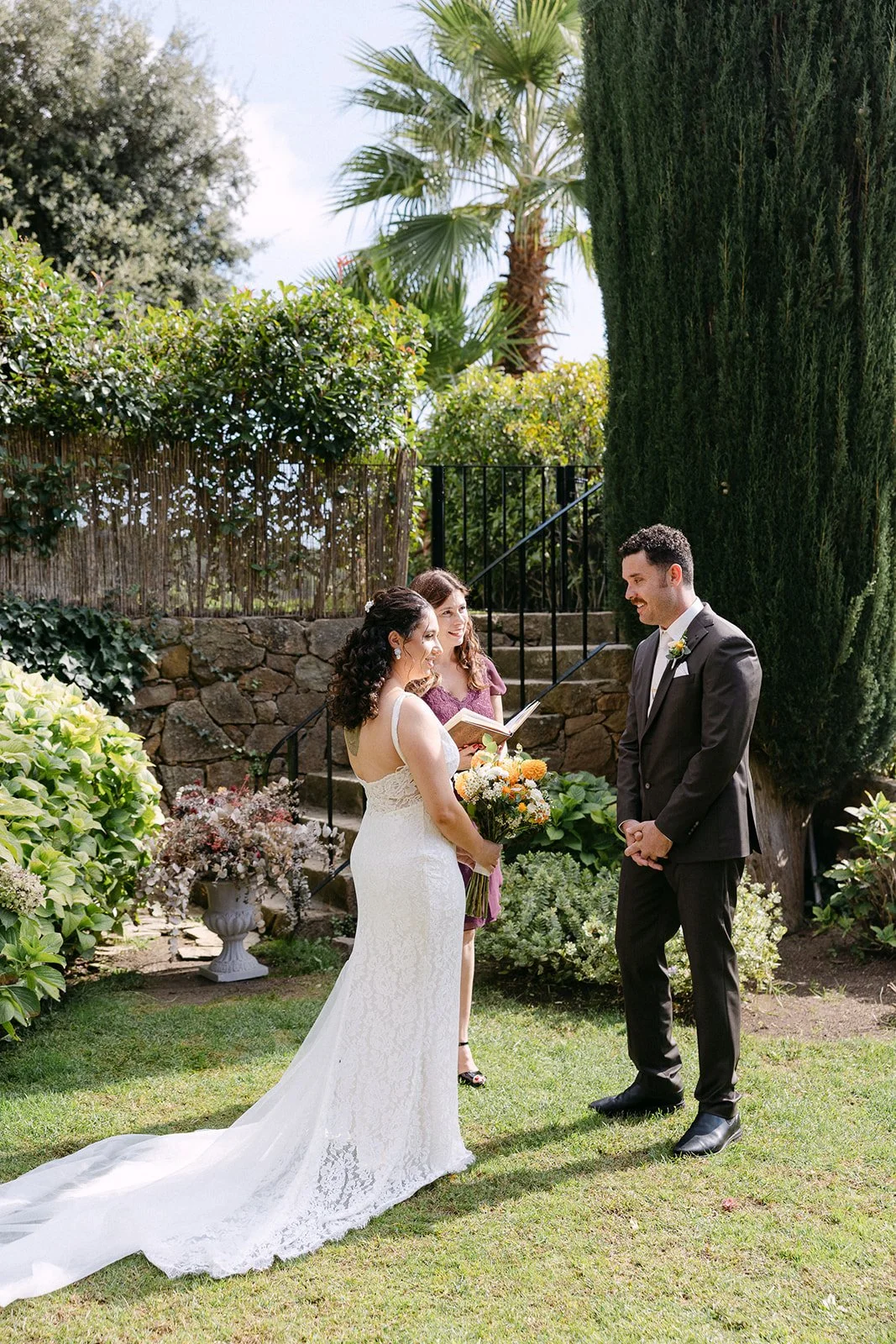 Bride and groom exchanging vows outside in a garden, with an officiant holding a book. The bride is in a white lace wedding dress holding a bouquet, and the groom is in a black suit. Lush greenery and stone steps are in the background.