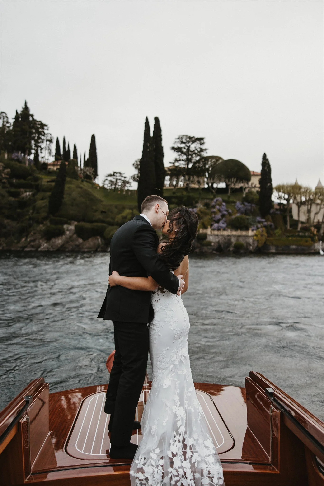 A couple dressed in wedding attire embracing on a boat on a river with a lush, hilly landscape and tall cypress trees in the background.