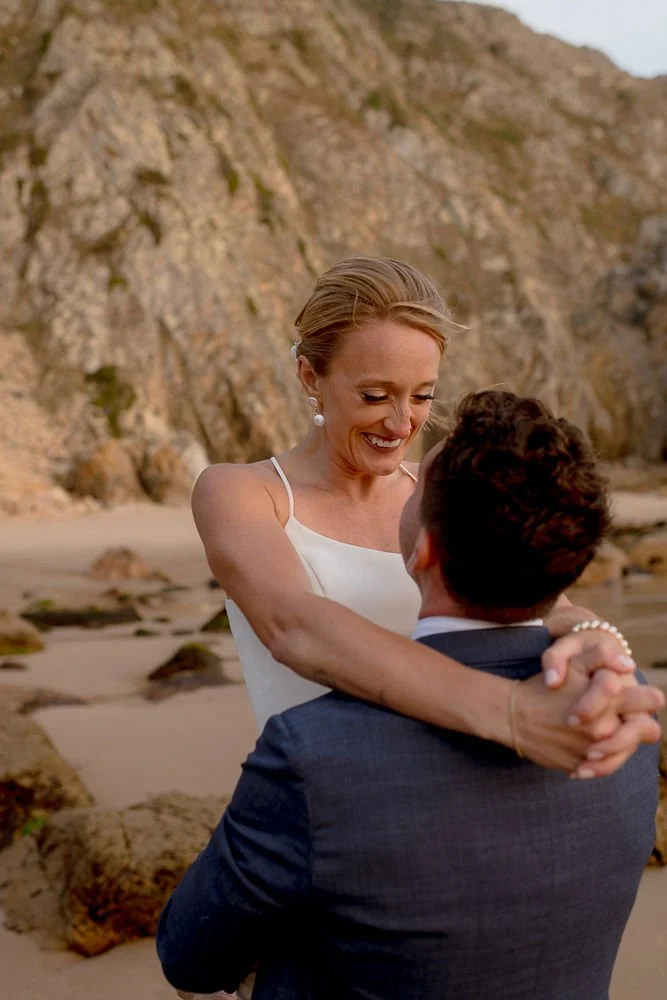 A woman in a white dress is embracing and smiling at a man in a suit on a beach with rocky cliffs in the background.