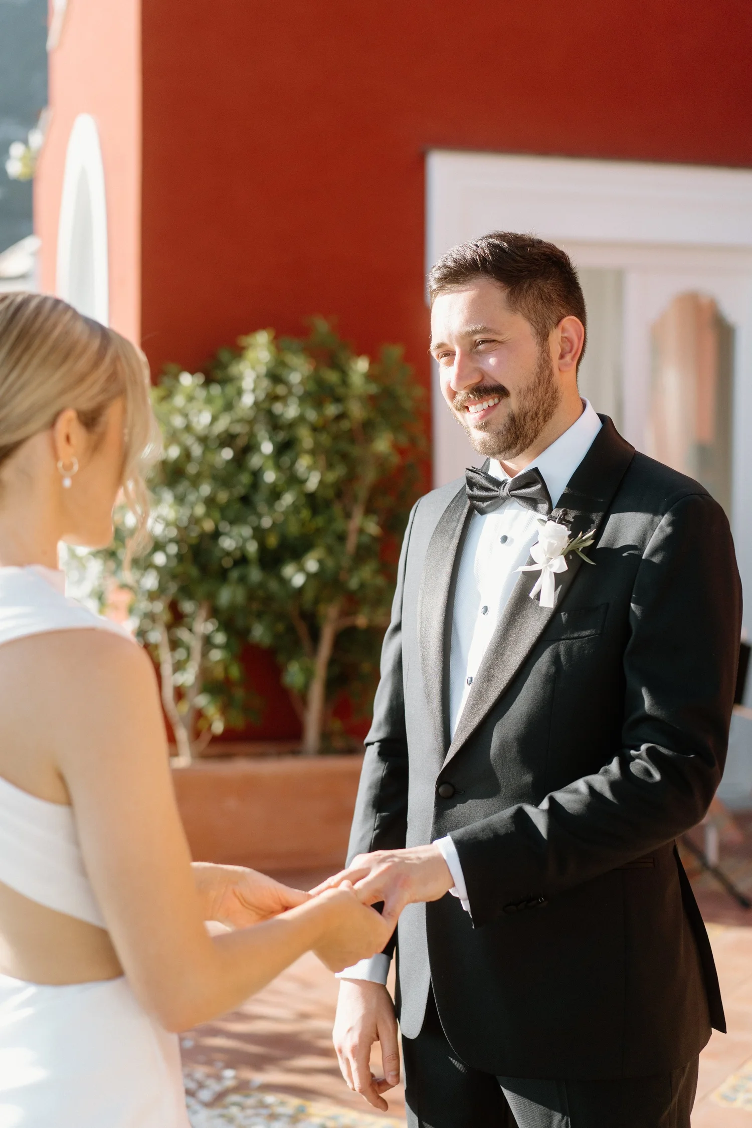 A man in a tuxedo smiling and holding hands with a woman in a white dress during a wedding ceremony outdoors.