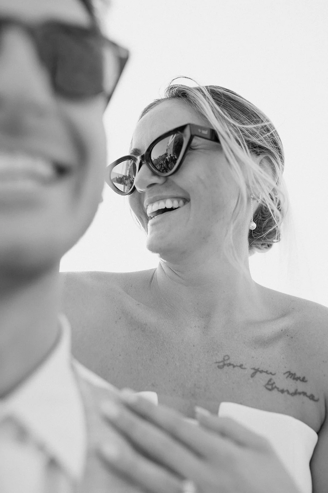 A black and white photograph of a woman and a child. The woman is smiling, wearing large sunglasses, and has a tattoo on her upper chest that says, "Love your more grandma." The child is slightly out of focus in the foreground.