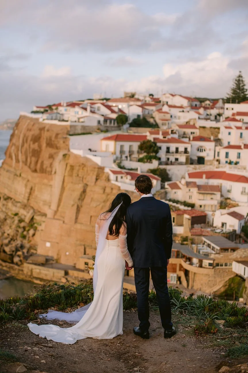 A bride and groom holding hands and facing a town with white buildings and red roofs, overlooking a cliff during sunset.