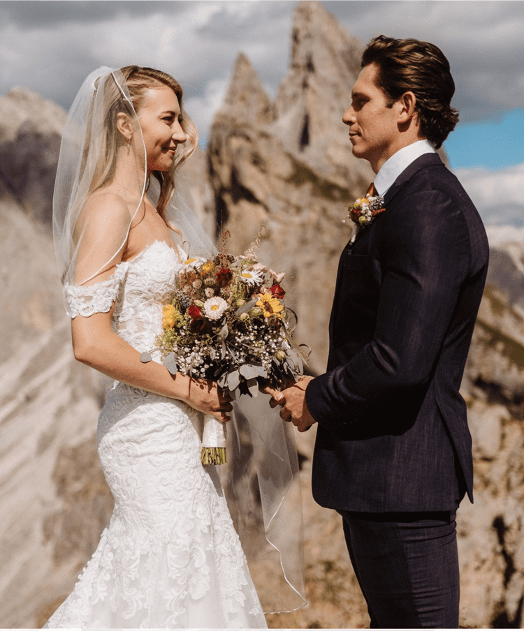 A bride and groom stand outdoors, holding hands and gazing at each other on their wedding day with rocky mountain peaks in the background.