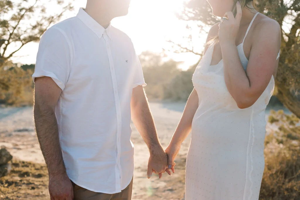 A couple holding hands outdoors, with the woman appearing pregnant, during sunset.