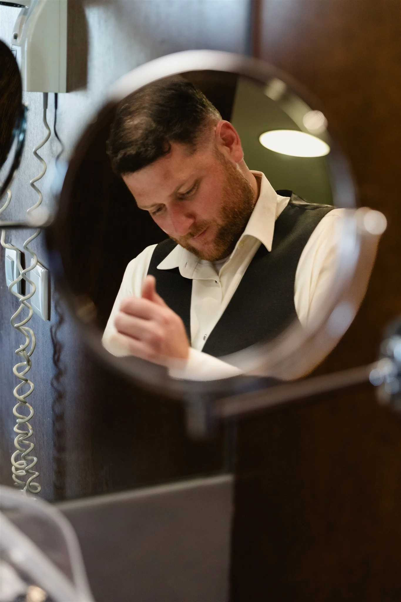 A man with a beard, wearing a white shirt and black vest, is looking down, captured through the reflection in a mirror.