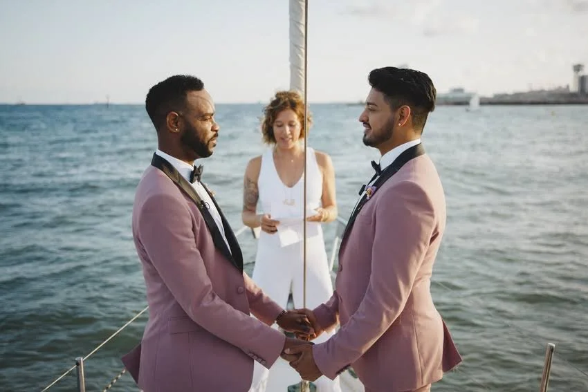 Two men in pink tuxedos holding hands during a wedding ceremony on a boat, with a female officiant in a white dress standing behind them, and water and city skyline in the background.